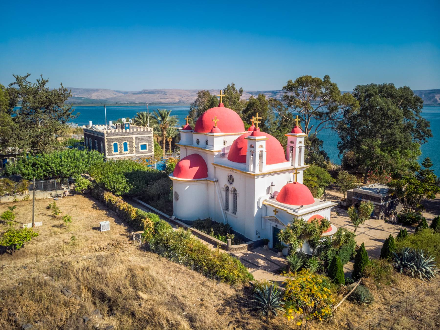 A white church with red domes near a body of water, surrounded by vegetation under a clear blue sky.