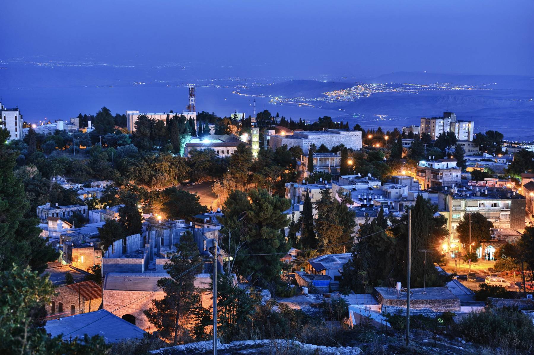 A panoramic nighttime view of a city with illuminated buildings and streets, overlooking a coastal area.