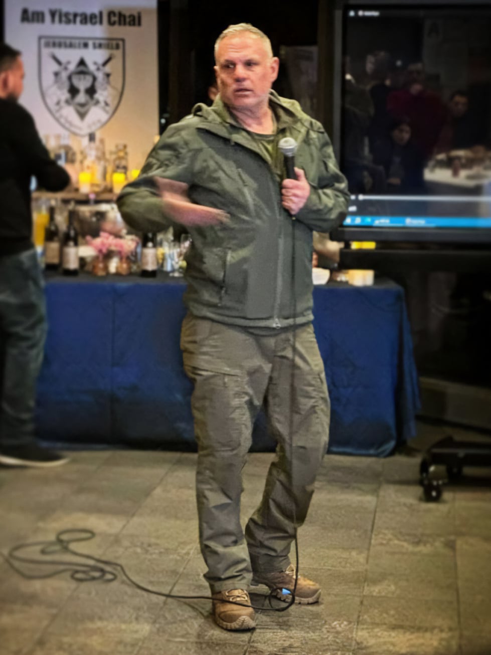 A man in military-style clothing speaks into a microphone indoors, with a table and a banner in the background. A microphone cord is visible at his feet.